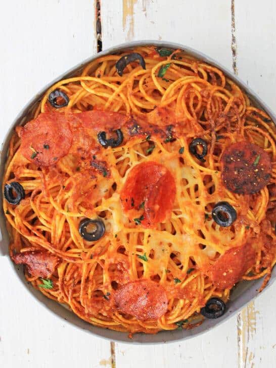 An air fryer loaded pizza pasta dish topped with melted cheese, pepperoni slices, and black olives, served in a round baking dish on a rustic white wooden table.