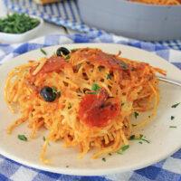 A plate of air fryer loaded pizza pasta topped with tomato sauce, olives, and chopped parsley, with a loaf of bread and a casserole dish in the background, on a blue and