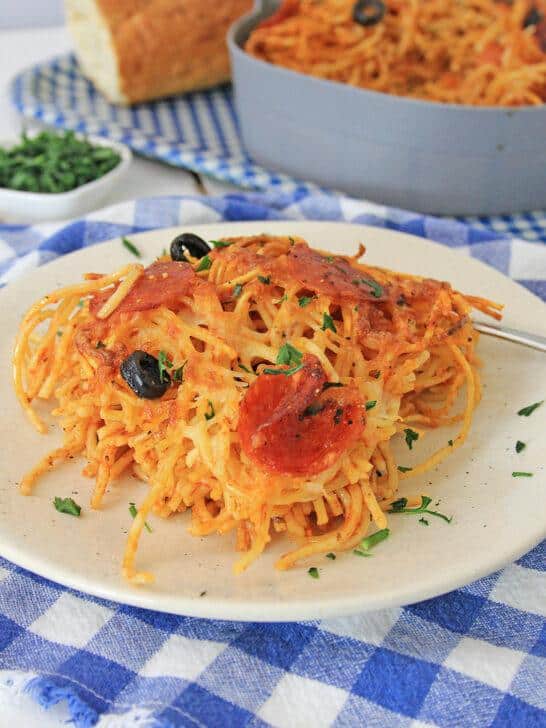 A plate of air fryer loaded pizza pasta topped with tomato sauce, olives, and chopped parsley, with a loaf of bread and a casserole dish in the background, on a blue and