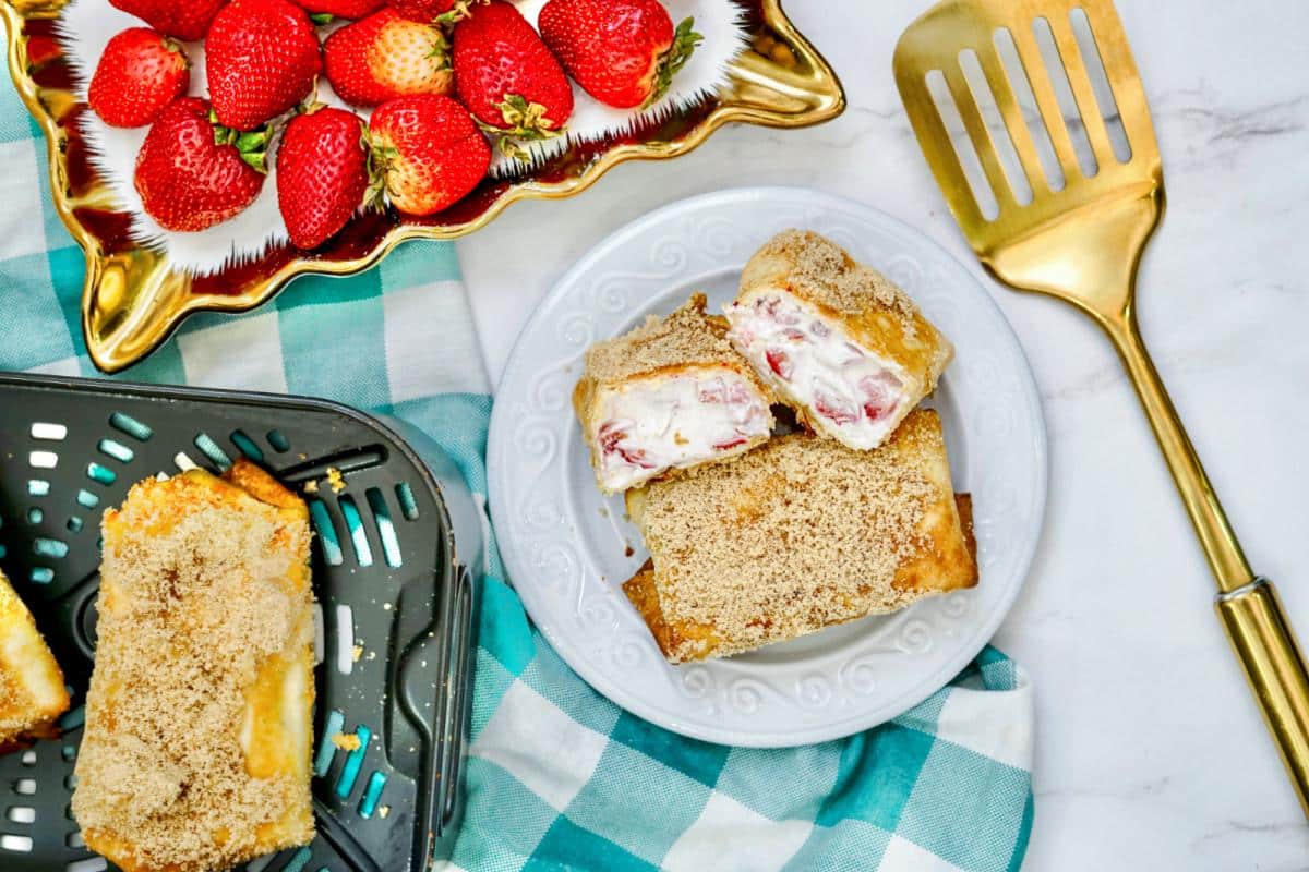 Top-down view of a plate with two slices of sweet strawberry cheesecake topped with crumbles, a golden spatula, and a bowl of strawberries on a marble surface.