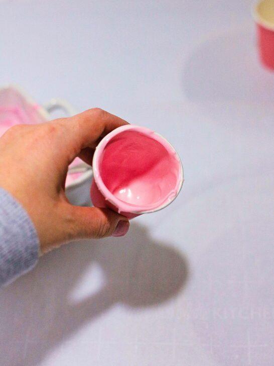 A person is holding a cup of pink icing, enjoying their Bailey’s Strawberry and Cream Cocoa Cups, a delectable Valentine's Day treat.