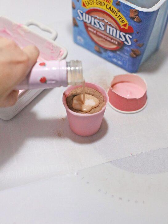 A hand delicately pouring Bailey's Strawberry and Cream Cocoa into a cup.