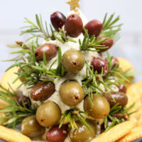 A holiday-themed appetizer shaped like a tree, crafted from garlic herb cheeses, green and black olives, and rosemary sprigs. It's topped with a star and surrounded by crackers on a white plate.