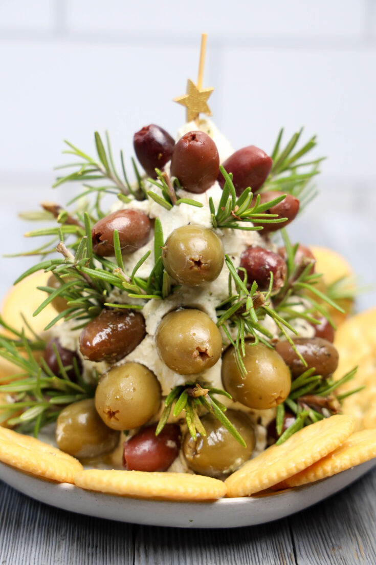 A holiday-themed appetizer shaped like a tree, crafted from garlic herb cheeses, green and black olives, and rosemary sprigs. It's topped with a star and surrounded by crackers on a white plate.