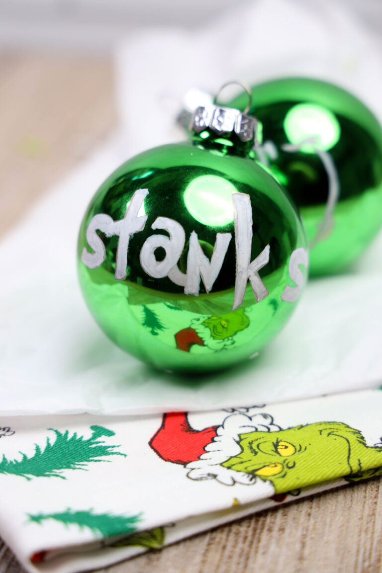 Grinch glass ornaments featuring the word "stank" are displayed on a napkin adorned with playful cartoon Grinch designs.