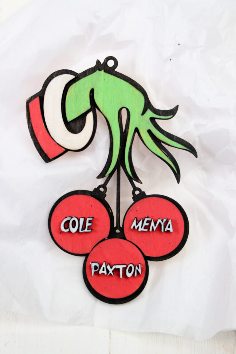 A Grinch wood ornament handily holds three red baubles labeled "Cole," "Menya," and "Paxton" against crumpled white paper.