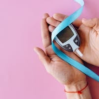 Hands holding a glucometer with a blue ribbon symbolizing diabetes awareness on a pink background.