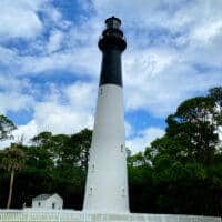 A black and white lighthouse stands tall with a surrounding white picket fence, green lawn, trees, and a partly cloudy sky in the background—one of the picturesque things to do in South Carolina.