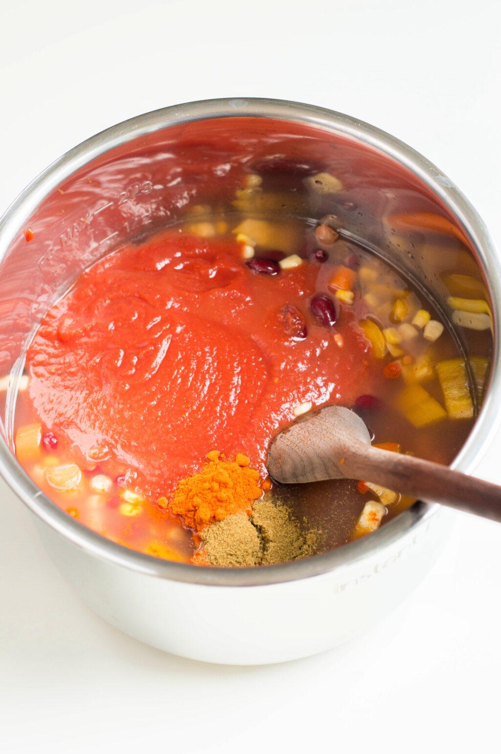 Bowl of vegetarian instant pot chili with a wooden spoon, featuring spices and corn kernels, on a white background.