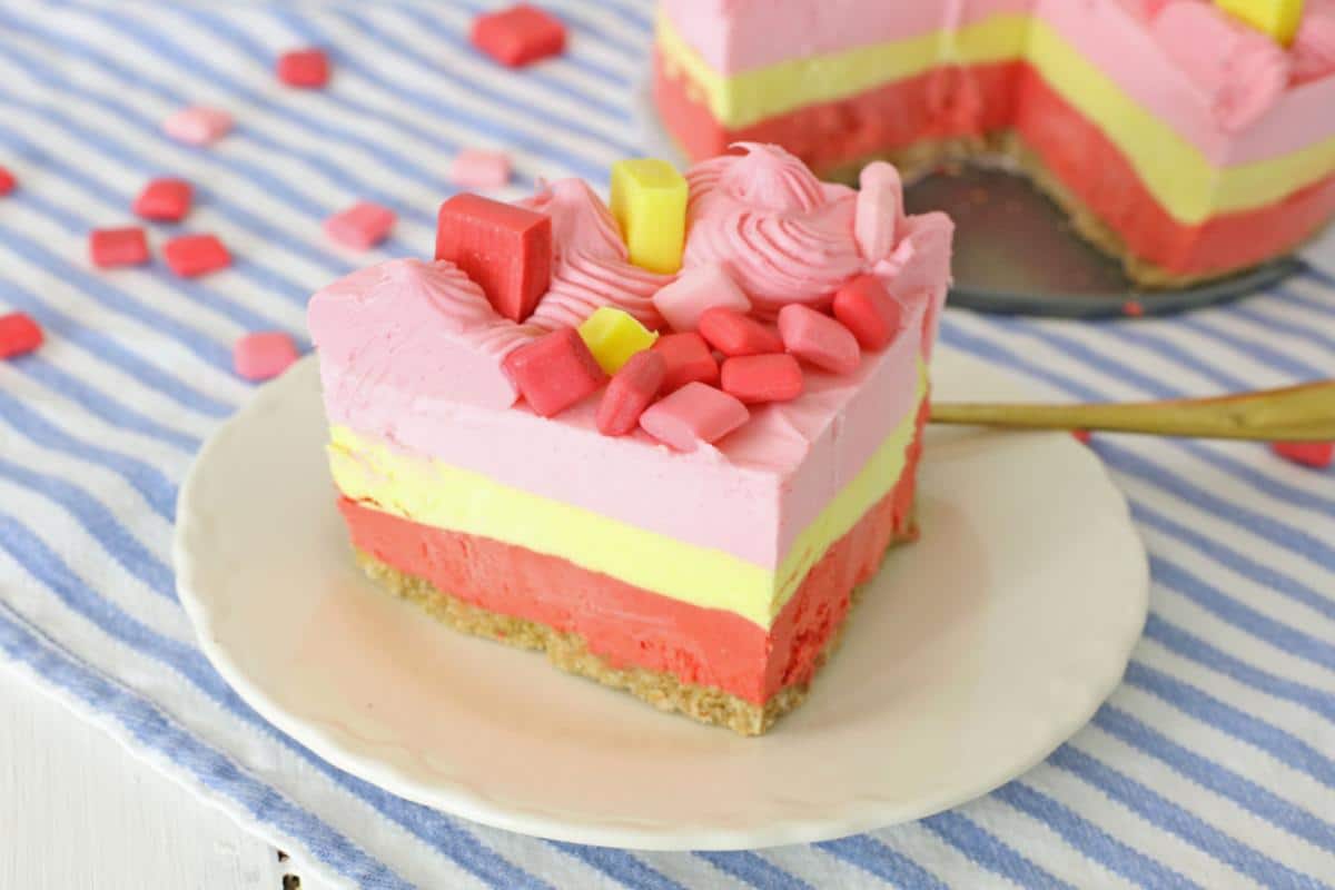 A slice of layered pink and yellow cheesecake topped with colorful candy, served on a white plate with scattered candies around on a striped tablecloth, presents a tempting dessert.