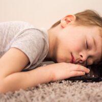 A child with autism is sleeping with their head resting on a laptop keyboard on a carpeted floor.