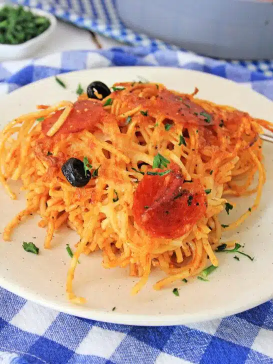 A plate of Air Fryer Loaded Pizza Pasta with melted cheese, sliced pepperoni, black olives, and garnished with chopped parsley, on a blue and white checkered tablecloth.