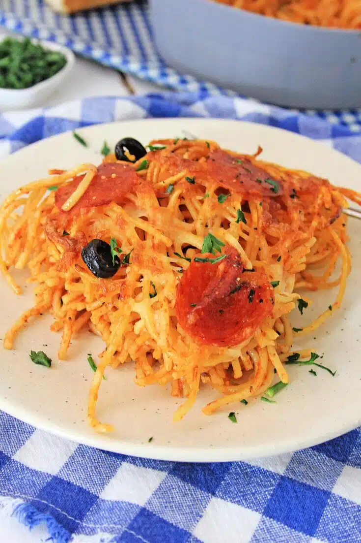 A plate of Air Fryer Loaded Pizza Pasta with melted cheese, sliced pepperoni, black olives, and garnished with chopped parsley, on a blue and white checkered tablecloth.