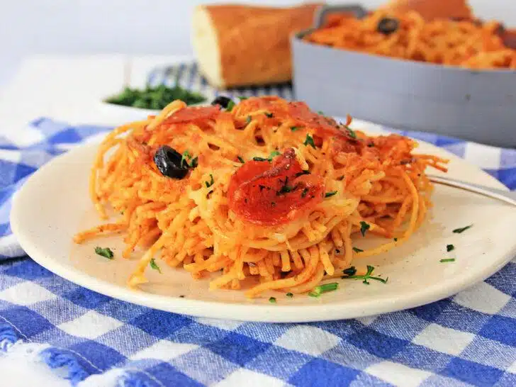 A plate of loaded pizza pasta with tomato sauce and olives, garnished with herbs, served on a blue and white checkered napkin.