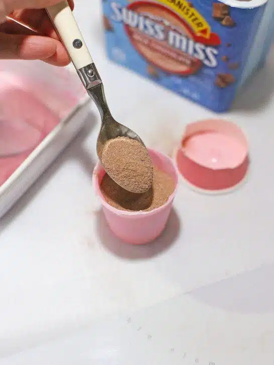 A person using a pink spoon to mix powdered sugar into a bowl of Bailey's Strawberry and Cream Cocoa Cups, preparing a Valentine's Day treat.