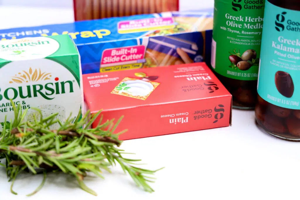 A selection of kitchen items, including Boursin cheese infused with garlic and herbs, plastic wrap, toothpicks, Good & Gather olive medleys from an ancient olive tree, and fresh rosemary beautifully arranged on a white surface.