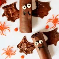 Two Halloween Bat Snack Cakes with chocolate rolls, adorned with candy eyes and bat wings made of chocolate, are surrounded by festive orange candy spiders and leaves.