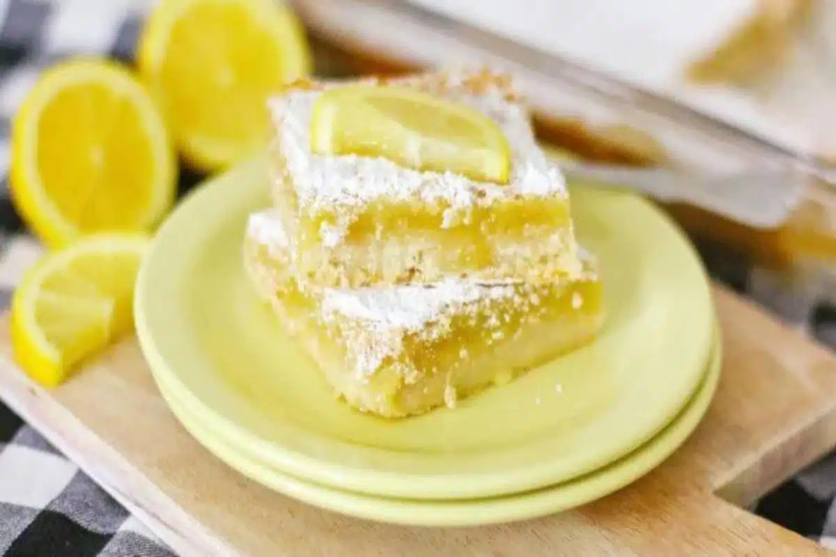 A slice of lemon bar topped with powdered sugar, served on a yellow plate with fresh lemon slices, against a checkered black and white cloth background. This sweet treat is perfect for dessert lovers.