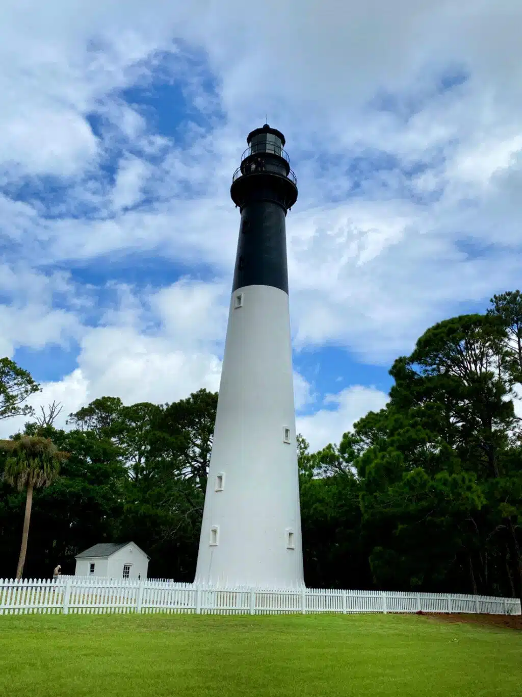 A black and white lighthouse stands tall with a surrounding white picket fence, green lawn, trees, and a partly cloudy sky in the background—one of the picturesque things to do in South Carolina.