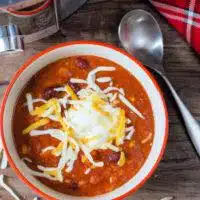 Bowl of Instant Pot Vegetarian Chili topped with shredded cheese and a dollop of sour cream, on a wooden table with a spoon and a checkered napkin on the side.