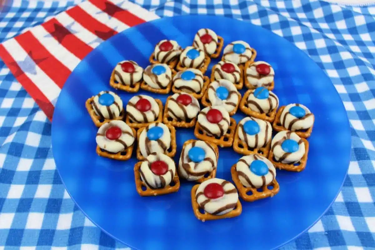 A blue plate with pretzel bites topped with melted chocolates and red, white, and blue candy, placed on a blue and white checkered tablecloth. An American flag-themed napkin in the background completes this fun patriotic recipe.