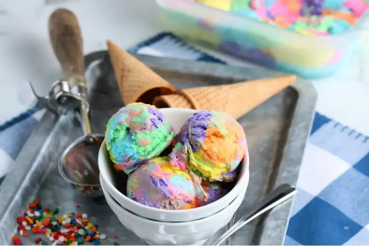 Three scoops of vibrant, multicolored ice cream served in a silver bowl with waffle cones and sprinkles on the side, creating a tempting dessert.